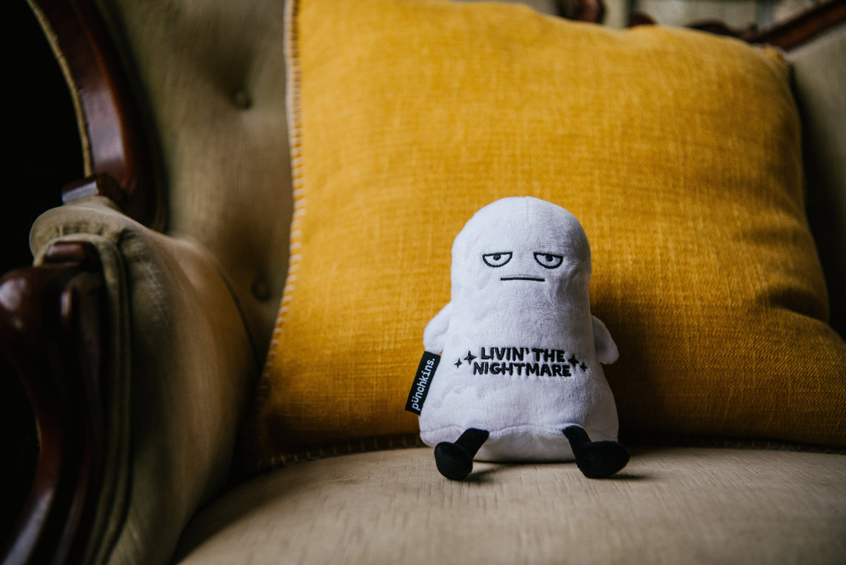Small grumpy ghost plush sitting on a mustard yellow pillow in a vintage armchair
